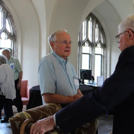 Two elderly men engaged in conversation in a room with arched windows and patterned chairs.
