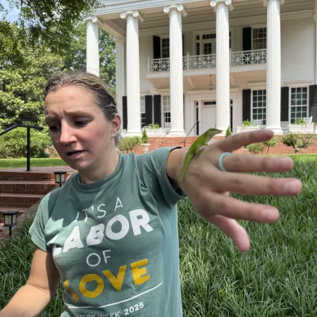 Woman with green shirt holding out her hand with a small green lizard outside a white columned house.