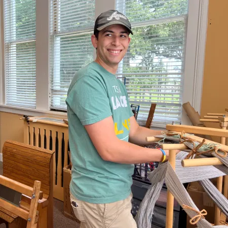 Young man smiling while weaving yarn on a wooden loom in a bright room with large windows.