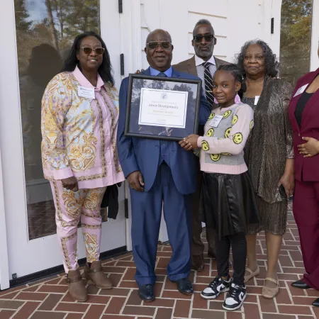 Group of six people standing outside, one man holding a framed certificate, smiling at the camera