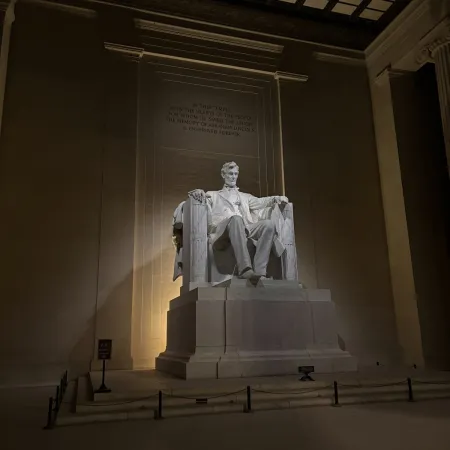 Illuminated statue of Abraham Lincoln seated in the Lincoln Memorial at night with engraved quote behind.