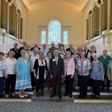 Group of elderly men and women posing inside a bright church with large organ pipes and a stained glass window.