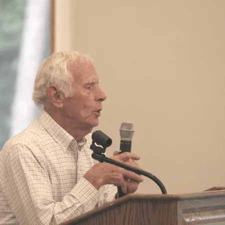Elderly man speaking into a microphone at a wooden podium during an indoor event.