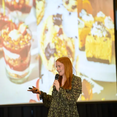 Young woman giving a presentation with a microphone in front of a dessert-themed slide with various sweets.