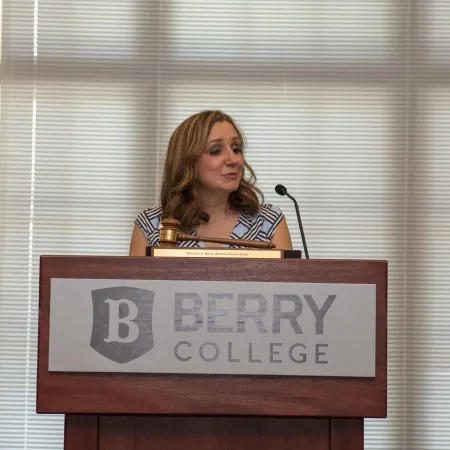 Woman speaking at Berry College podium with microphone and gavel in a conference room.