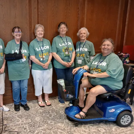 Six elderly women wearing matching green shirts with Labor of Love text, standing and sitting in a wood-paneled room.