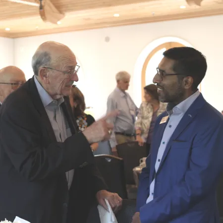 Two men in business attire having a conversation at a professional indoor event with guests in the background.