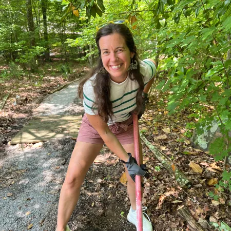 Smiling woman gardening with a shovel on a forest path surrounded by green trees and leaves.