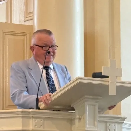 Man in a light blue suit speaking at a church pulpit with a cross in the background
