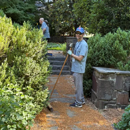 Person raking leaves on gravel path in lush garden with greenery and stone steps in background