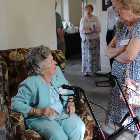 Two elderly women conversing in a living room, with a walker and additional people in the background.