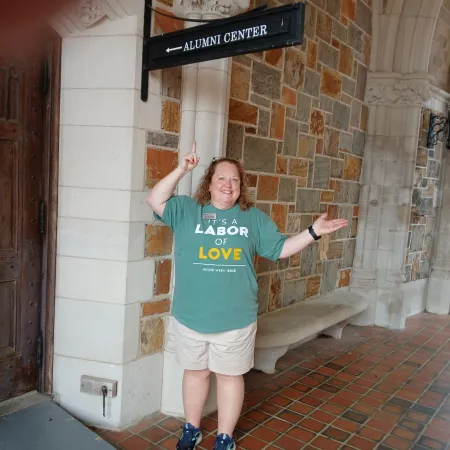 Smiling woman in green 'Labor of Love' shirt pointing to Alumni Center sign in stone hallway with arched ceiling.