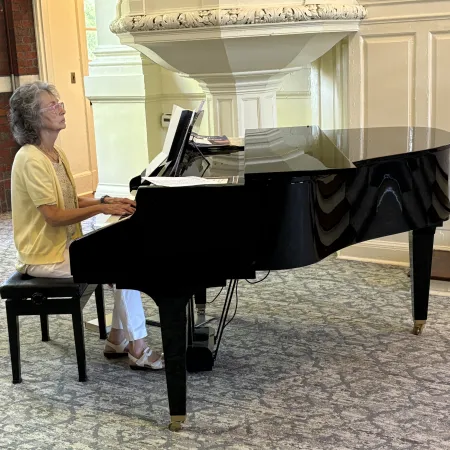 Older woman playing a black grand piano in a bright room with brick and white walls and patterned carpet.