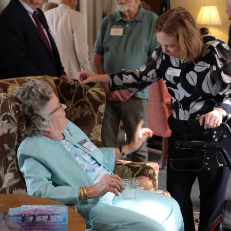 Two women engaged in conversation, one seated with a walker, in a cozy living room setting with three men in background.