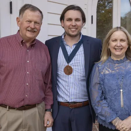 Three people, including a young man with a medal, standing together outdoors near a building entrance.