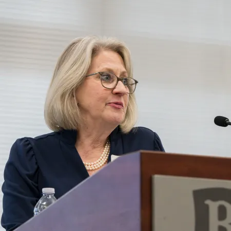 Mature woman speaking at podium with microphone in a professional setting, wearing glasses and pearl necklace.