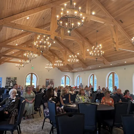 Audience attentively seated in a large, wood-beamed conference room with arched windows and chandeliers.