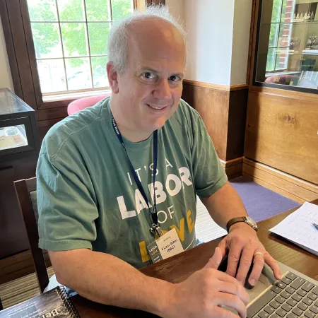 Man wearing a green shirt sits at a wooden desk using a laptop in a room with large windows and display cabinets.