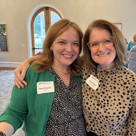 Two smiling women wearing name tags at an indoor social event with people in the background