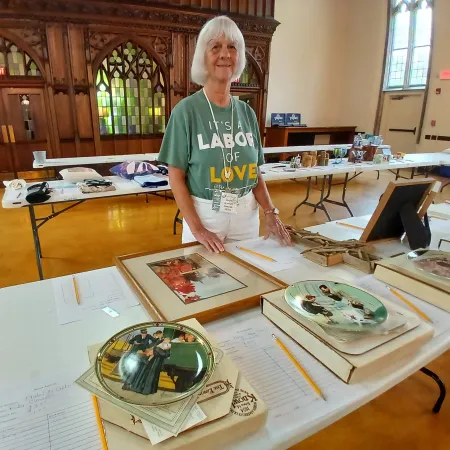 Woman standing behind table with vintage photos, plates, and memorabilia displayed for an event in a large room.