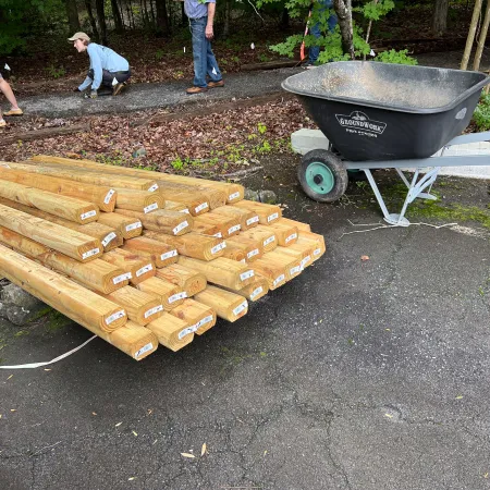 Stack of wooden posts beside a wheelbarrow on an asphalt path with people working in the background