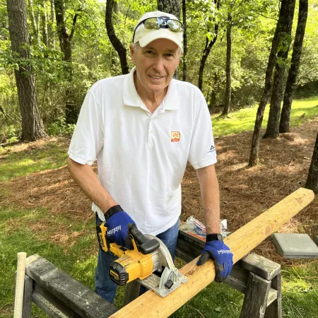 Man wearing white shirt and gloves cutting wooden beam with circular saw outdoors in forested area