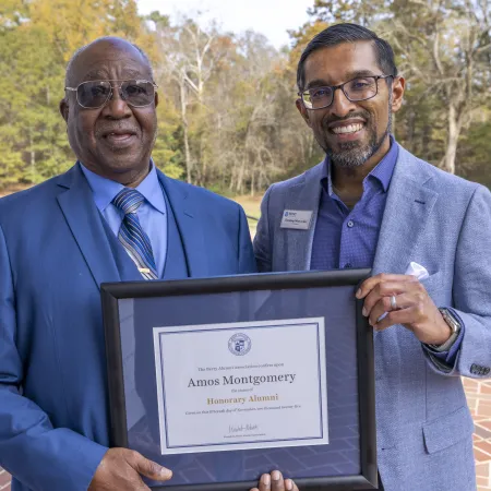Two men outdoors holding a framed honorary alumni certificate for Amos Montgomery on a sunny day