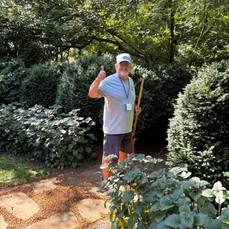 Man wearing a white cap and shirt holding a walking stick in a lush green garden pathway on a sunny day