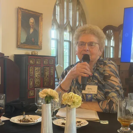 Elderly woman speaking into a microphone at a table with flowers and plates in a room with stained glass windows and a portrait.