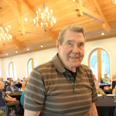 Elderly man in striped shirt and beige pants smiling at a social event in a warmly lit wooden hall.