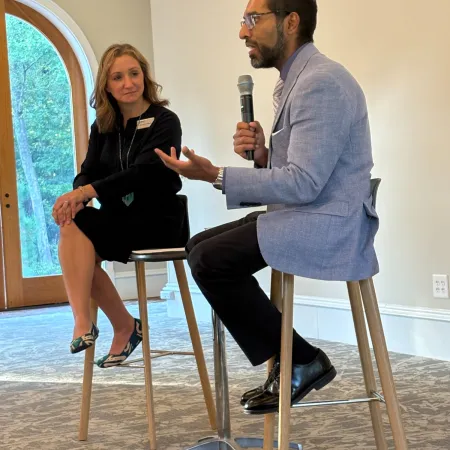 Man in blazer interviews woman at a professional event with microphone on high stools indoors near a window.