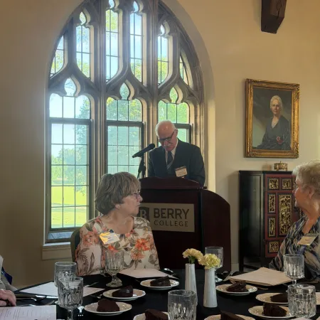 Man speaking at a Berry College podium in a sunlit room with large arched windows and seated attendees.