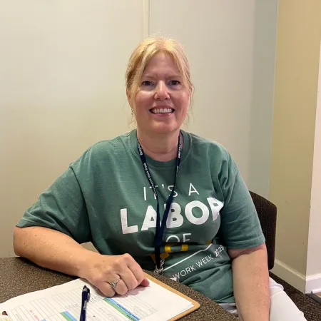 Smiling woman wearing green Labor Day 2023 t-shirt sitting at a desk with a clipboard and pen in an office.
