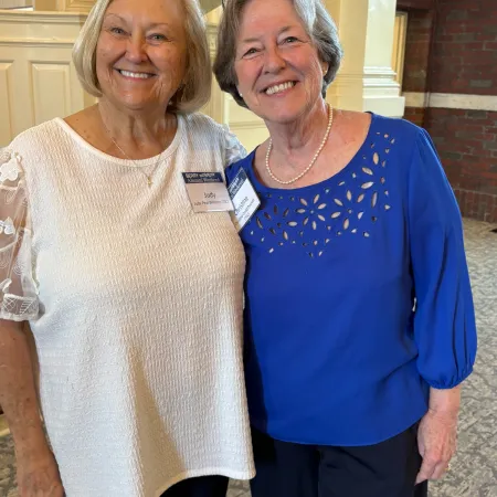 Two elderly women smiling indoors, one in a blue blouse and the other in a white textured top.