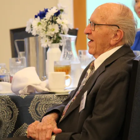 Elderly man in suit sitting relaxed with hands clasped at formal event with table setting and flowers in background