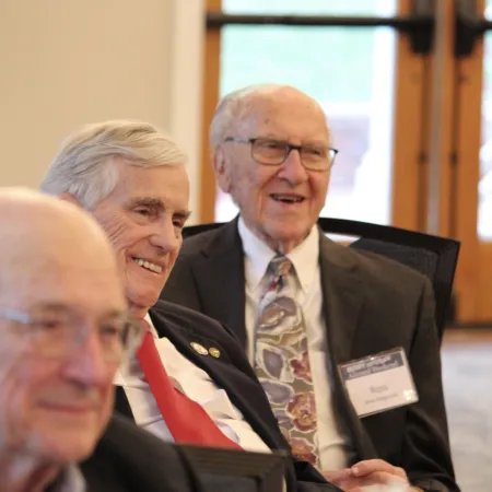 Three elderly men in suits smiling and socializing indoors at a formal event or conference.