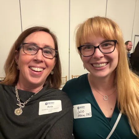 Two smiling women wearing glasses and name tags at an indoor event with other attendees in the background.