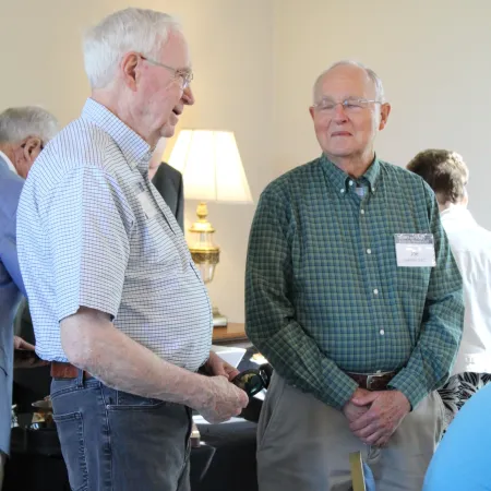Two elderly men engaged in conversation at an indoor event with name tags and casual attire.