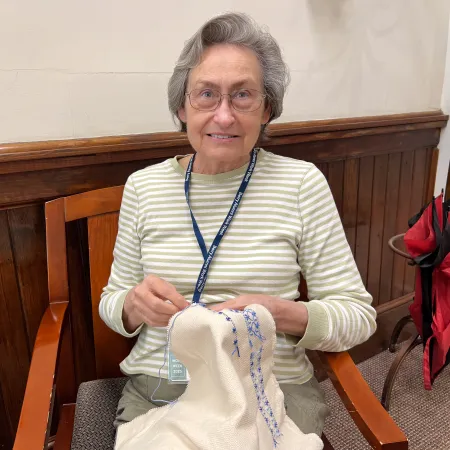 Smiling elderly woman wearing glasses seated and stitching blue embroidery on white fabric indoors.