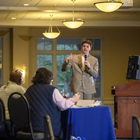 Young man in a suit speaking with a microphone to seated audience in a well-lit room with large windows.