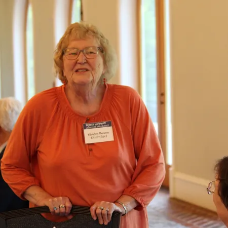 Elderly woman in orange shirt with glasses smiling at a social event indoors with natural light.