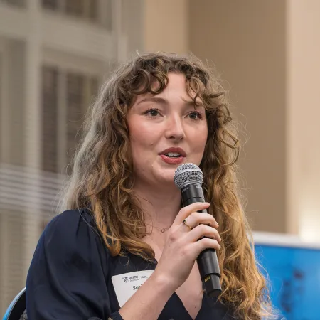 Woman with curly hair speaking into microphone while holding white roses wrapped in plastic.