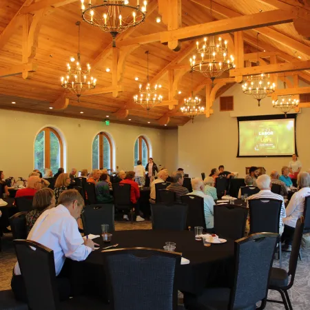 Community meeting with seniors seated around tables in a spacious room with wooden ceiling and chandeliers.