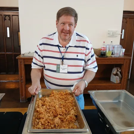 Man serving a tray of cooked rice at a buffet table in a wooden-paneled room