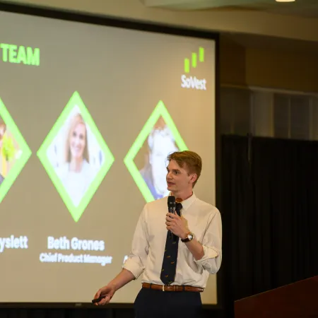 Man in white shirt and tie presenting company team slide with photos and roles at a conference or meeting.