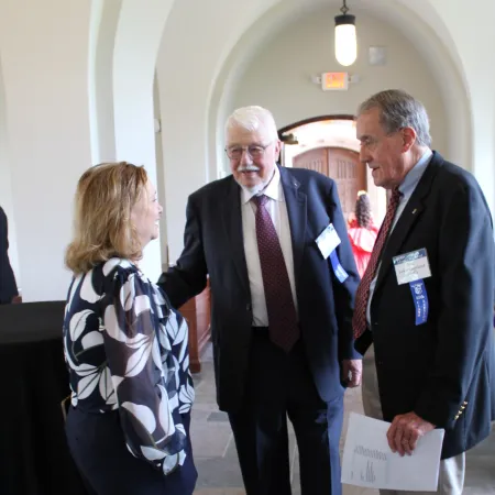 Three professionals in business attire conversing in a bright hallway with arched ceilings and tiled floor.