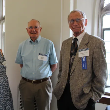 Three older adults with name tags and ribbons standing in a well-lit room with arched windows and office furniture.