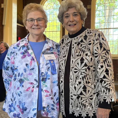 Two elderly women smiling indoors, one wearing a floral jacket and glasses, the other in a lace-patterned cardigan.