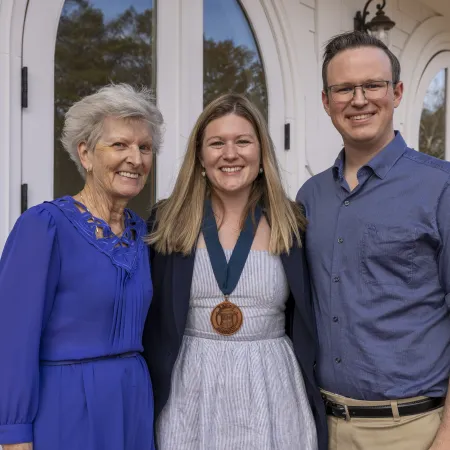 Three people smiling outdoors by a white building, young woman wearing a medal, flanked by an elderly woman and younger man.