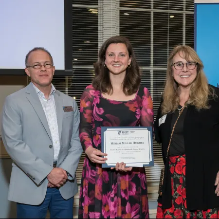Three people standing indoors; center woman holds certificate; Berry Alumni banner in background.